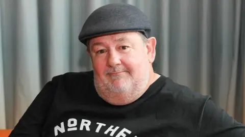 BBC Johnny Vegas wearing a dark flat cap and a black T-shirt with the word “NORTHERN” printed in bold white letters across the chest. The background consists of grey curtains.