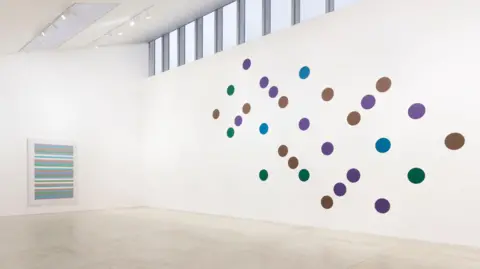 Turner Contemporary A gallery room with white walls and a row of windows near the roof. On one wall is a large painting consisting of multiple spots in purple, green and brown, seemingly arranged at random but somehow also with a sense of order. On the left is a painting comprised of several horizontal lines in green, pink, blue, white and some other colours.