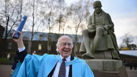 Getty Images Prof Higgs is looking very happy. He is wearing a pale blue graduation gown and is holding a scroll in the air. There is a statue behind him.