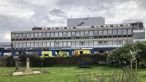 David Robinson / Geograph A long, wide building with numerous windows stands in the centre, with ambulances outside an entrance, a sign reads 'Darlington Memorial Hospital'. A park sits in front. 