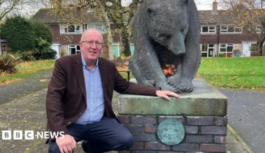 Stephen Fenton kneels beside a large bronze bear sculpture mounted on a brick and stone pedestal in an outdoor setting. The bear is positioned with its head lowered and front paws together, holding several apples. The pedestal features a circular plaque with an engraved design.