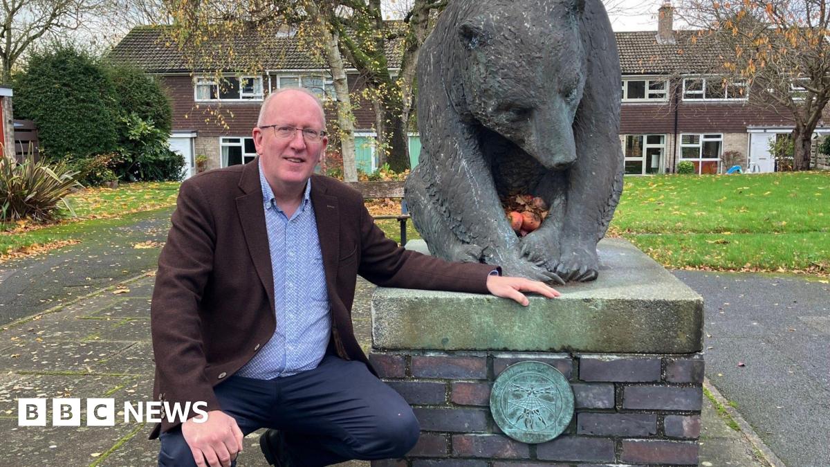 Stephen Fenton kneels beside a large bronze bear sculpture mounted on a brick and stone pedestal in an outdoor setting. The bear is positioned with its head lowered and front paws together, holding several apples. The pedestal features a circular plaque with an engraved design.