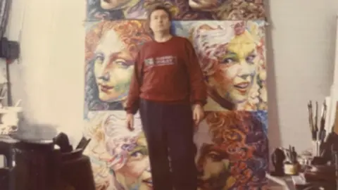 Henry Orlik An old and slightly grainy photo of Henry Orlik standing in front of a series of canvases, a couple of which are portraits of Marilyn Monroe in a brightly coloured style, stacked up against a wall.