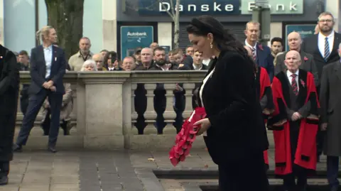 Emma Little Pengelly is holding a poppy wreath. Her head is bent. Crowds of people are watching.