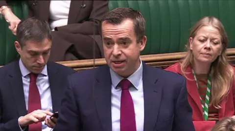 Parliament TV Calum Miller MP stands to speak in parliament. He has neat, dark hair and wears a dark suit, light shirt and magenta tie.