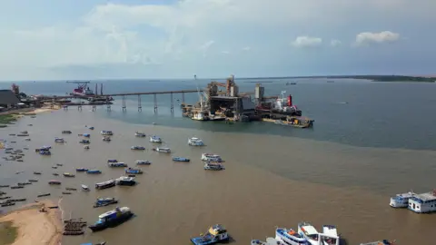 BBC / Tony Jolliffe A wide view of a port at the mouth of the Tajapos River in the Amazon river. You see a terminal and an industrial complex which is connected by a bridge. A number of ships and boats are in the water which looks brown close to the shore and blue away from the shore.  