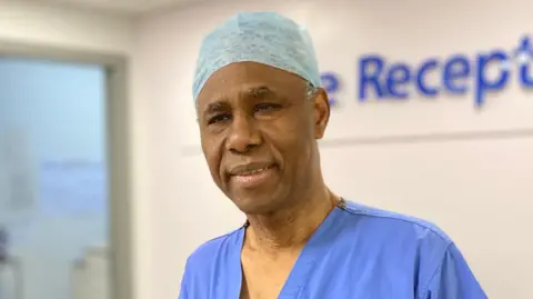 BBC Mr Akomolafe is stood in front of a hospital reception desk.  He is wearing theatre scrubs, which includes a light blue surgical cap and blue top.
