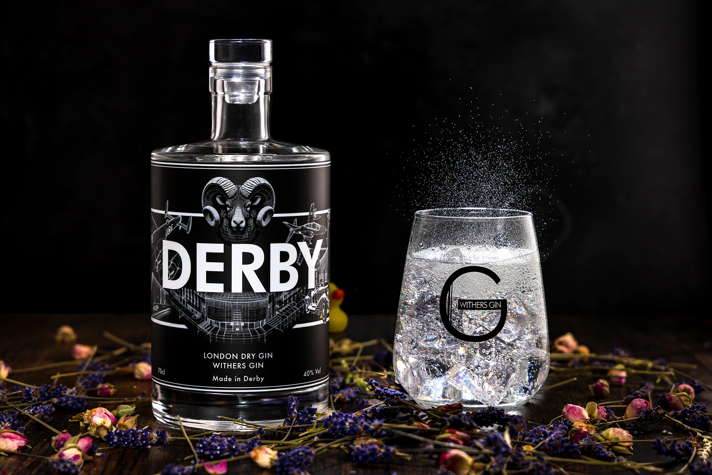 A bottle of Derby London Dry Gin next to a sparkling gin and tonic in a branded glass, surrounded by dried flowers on a dark background