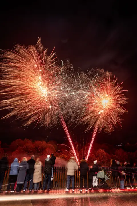 Duncan Loraine Brilliant red fireworks burst across a dark night sky, creating dazzling trails of light and sparks. A group of spectators stands in the foreground, watching the display from behind a railing.