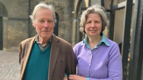A woman with short grey hair on the right wearing a button-up lilac cardigan and a blue shirt underneath and linking her arm with a man wearing a brown blazer, teal v jumper and a green shirt. He has short white hair.