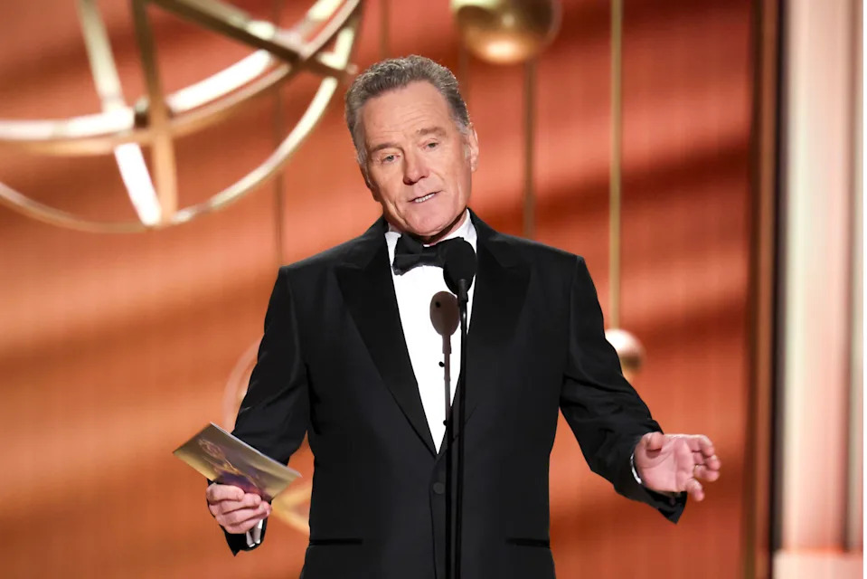 Bryan Cranston in a tuxedo speaks at an awards ceremony, holding an envelope. Large decorative element in background.