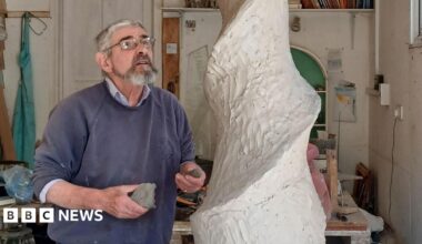 John working on the sculpture. He is looking up at a spiral made out of clay. He is wearing a blue jumper and glasses. He is in a studio.