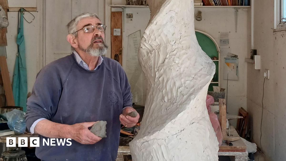 John working on the sculpture. He is looking up at a spiral made out of clay. He is wearing a blue jumper and glasses. He is in a studio.