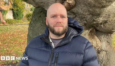 Pete White is an RAF veteran who left the armed forces in 2017. He is bald with a beard and is wearing a puffer jacket and standing in front of a tree, looking into the camera. There is a grassed area behind him.