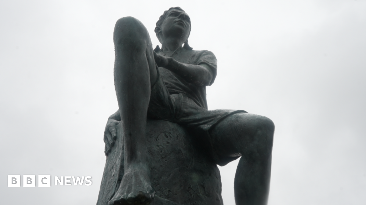 Zeb Soanes, wearing a blue shirt, and sculptor Ian Rank-Broadley, wearing a white jacket, stand either side of an alabaster sculpture of Benjamin Britten as a boy