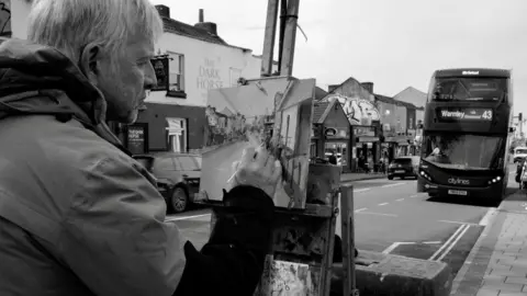 Bernard Bryden A black and white photo of artist Peter Brown painting a picture of a street. He is holding the paintbrush to the canvas and looking at his work. A bus heading for Warmley can be seen driving on the road ahead of him, and a pub and other businesses can be seen across the road. The photo is taken over Peter's shoulder, so only half his face can be seen.