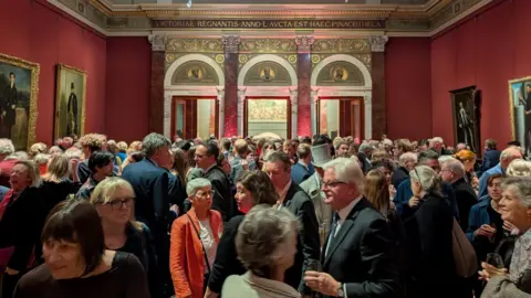 Derby Museums Trust Crowd of people looking at paintings in a large red room