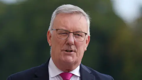 PA Media Mike Nesbitt looks to the right of the camera. He has short grey hair and wears clear glasses with black frames, as well ads a navy suit, white shirt and pink tie. 