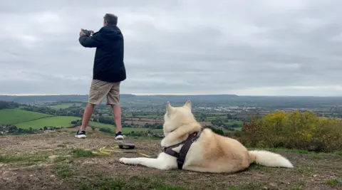 A white husky is lying on a hill overlooking a valley. The husky is looking to a man who is standing with his back to the camera.
