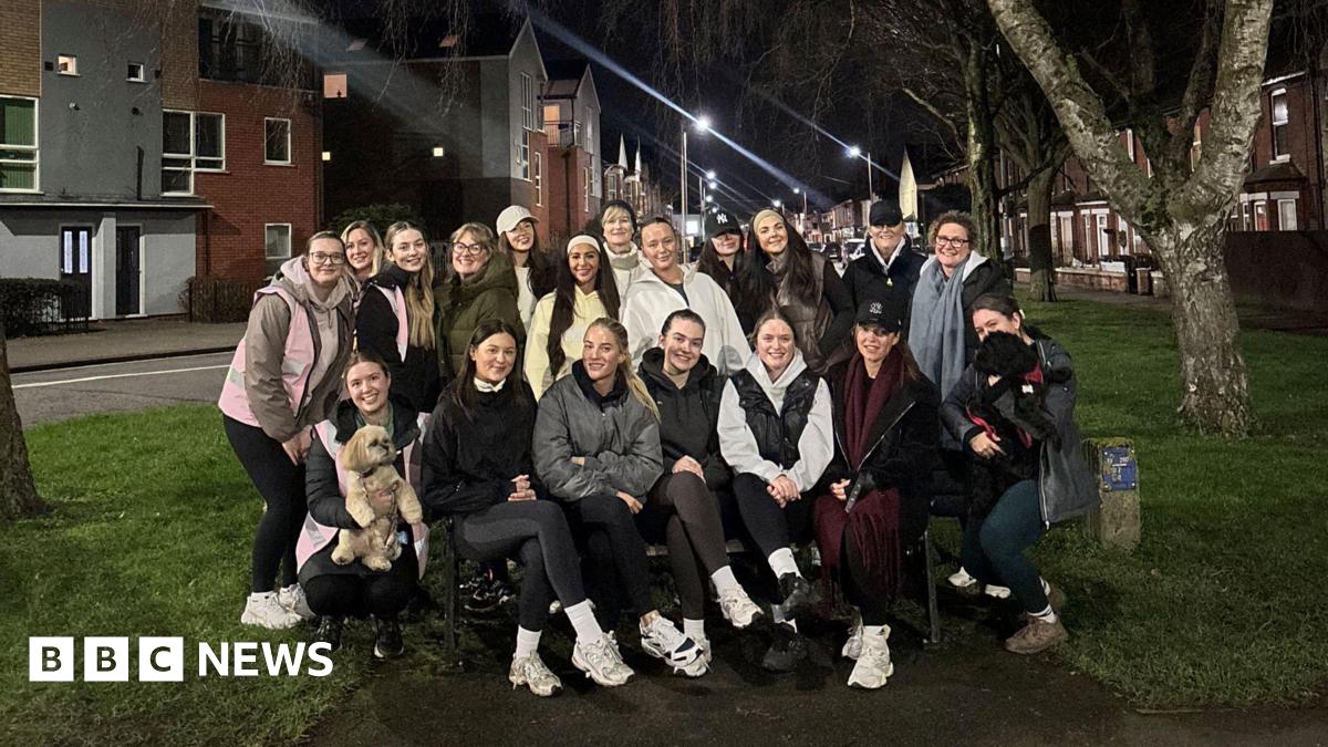 A group of girls, some sitting on an outdoor bench and some standing up, smiling at the camera. It is night time.