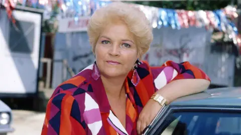 Pam St Clement as Pat Butcher in 1992 in a bright red, purple and orange blouse with large purple earrings, and leaning on a car roof