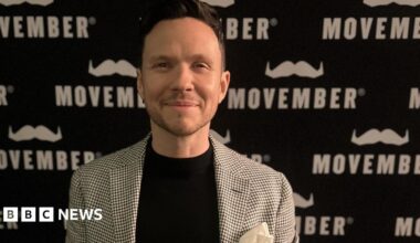 Bill Brewer, who is wearing a chequered blazer and black T-shirt, is standing in front of a black and white board with the Movember logo.