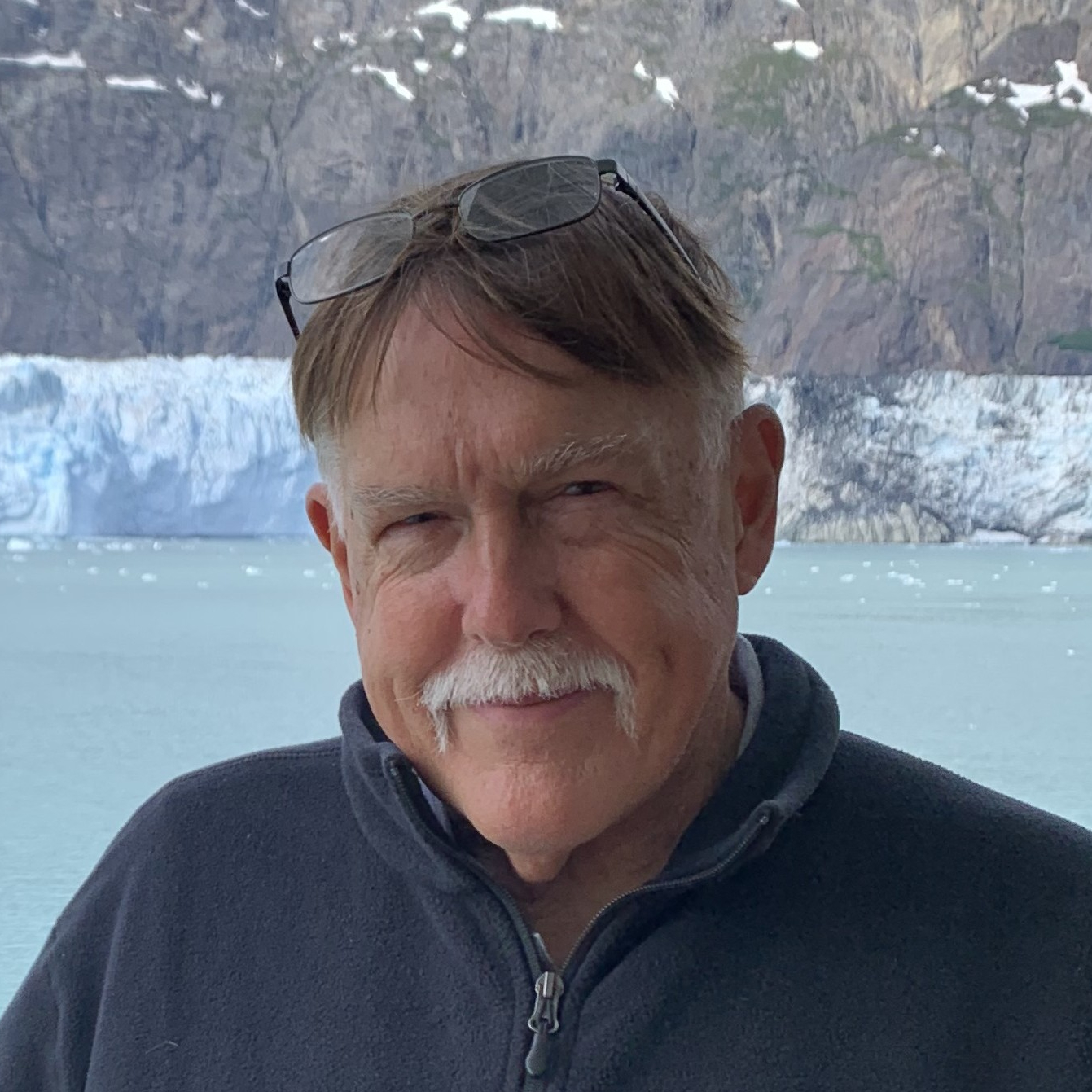 A man with a white moustache and graying hair with glasses on top of his head and a navy jacket stands in front of a frozen lake with snow in the background