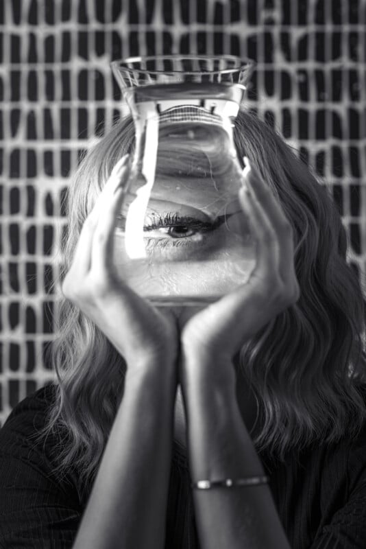 A person holds a glass of water in front of their face, distorting the appearance of their eye behind the glass. The image is in black and white, with a patterned background of wavy vertical lines.