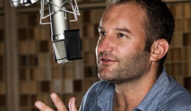 David Greene speaks into a large studio microphone, gesturing with one hand as he talks. He is seated in a recording booth with a blurred background of wooden acoustic panels.