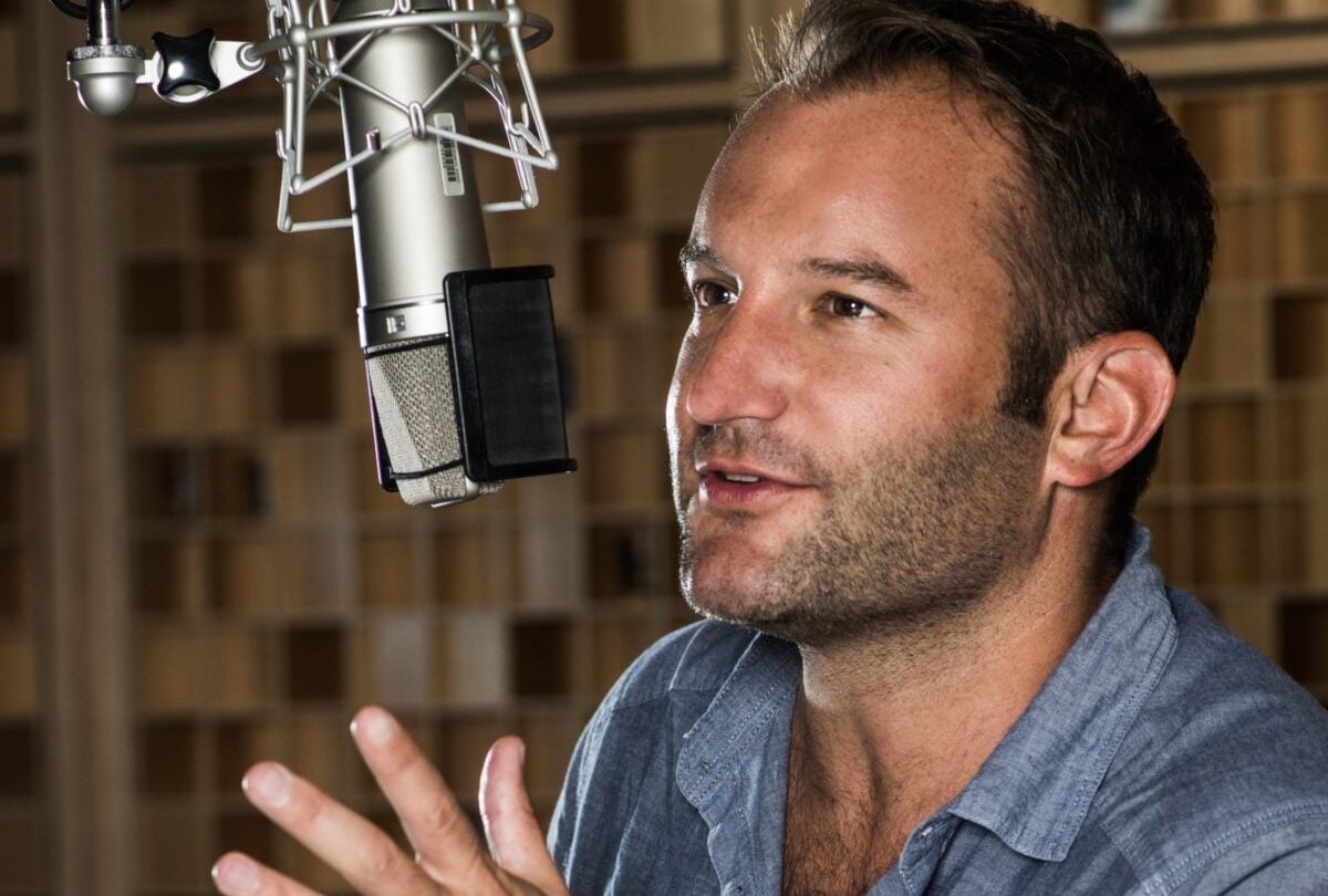 David Greene speaks into a large studio microphone, gesturing with one hand as he talks. He is seated in a recording booth with a blurred background of wooden acoustic panels.