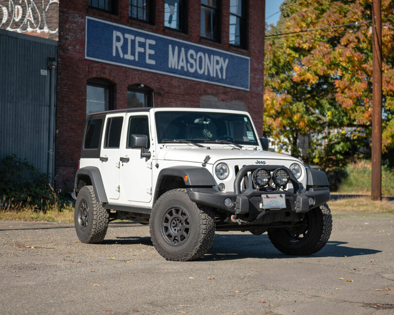A white Jeep Wrangler is parked on a paved lot in front of a brick building with a "Rife Masonry" sign.