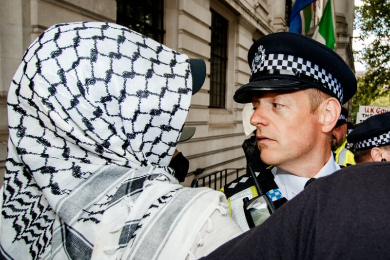 A protester wearing a keffiyeh faces a police officer in uniform at close range during a demonstration outside a government building. The scene appears tense, with both individuals making direct eye contact.