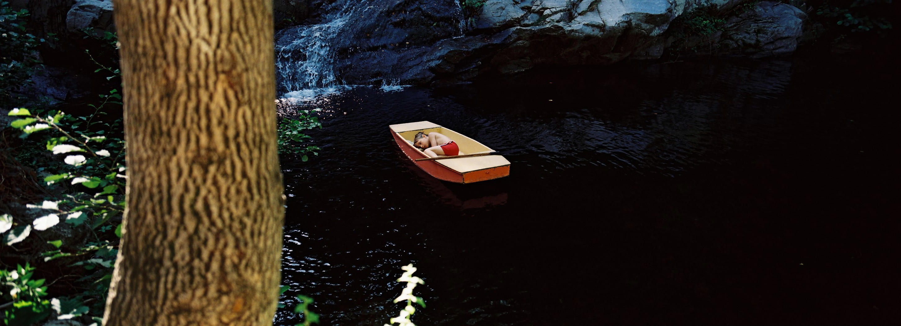 A serene scene featuring a small orange boat on dark waters, with a person reclining inside, surrounded by lush greenery and rocks