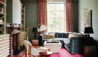 A living room with teal walls, a bright pink rug, and a teal velvet corner sofa in front of a large window.