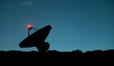 A dark silhouette of a satellite dish tilts to the left next to the dark horizon with a dark blue night sky in the background