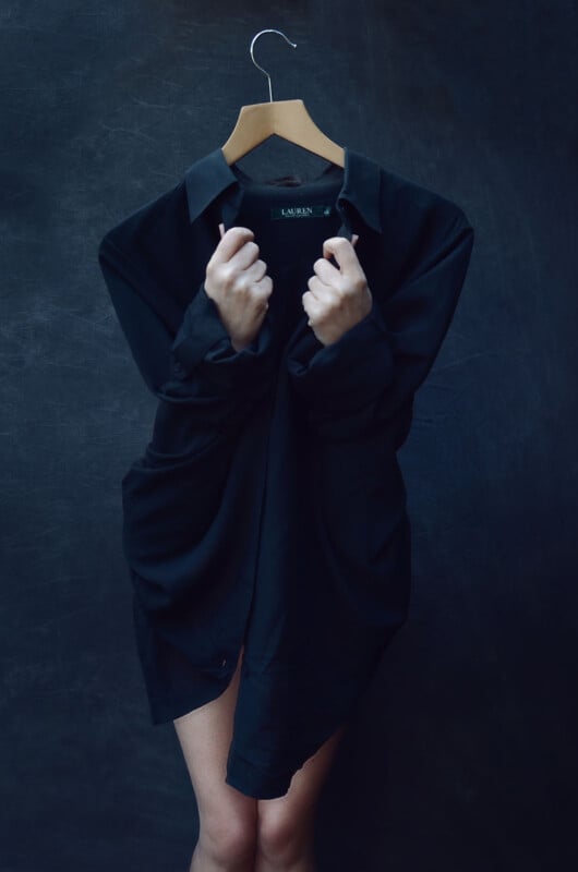 A person holds a black shirt on a hanger in front of their body, with only their bare legs and hands visible, against a dark background. The shirt covers their torso and face, creating a surreal, playful effect.