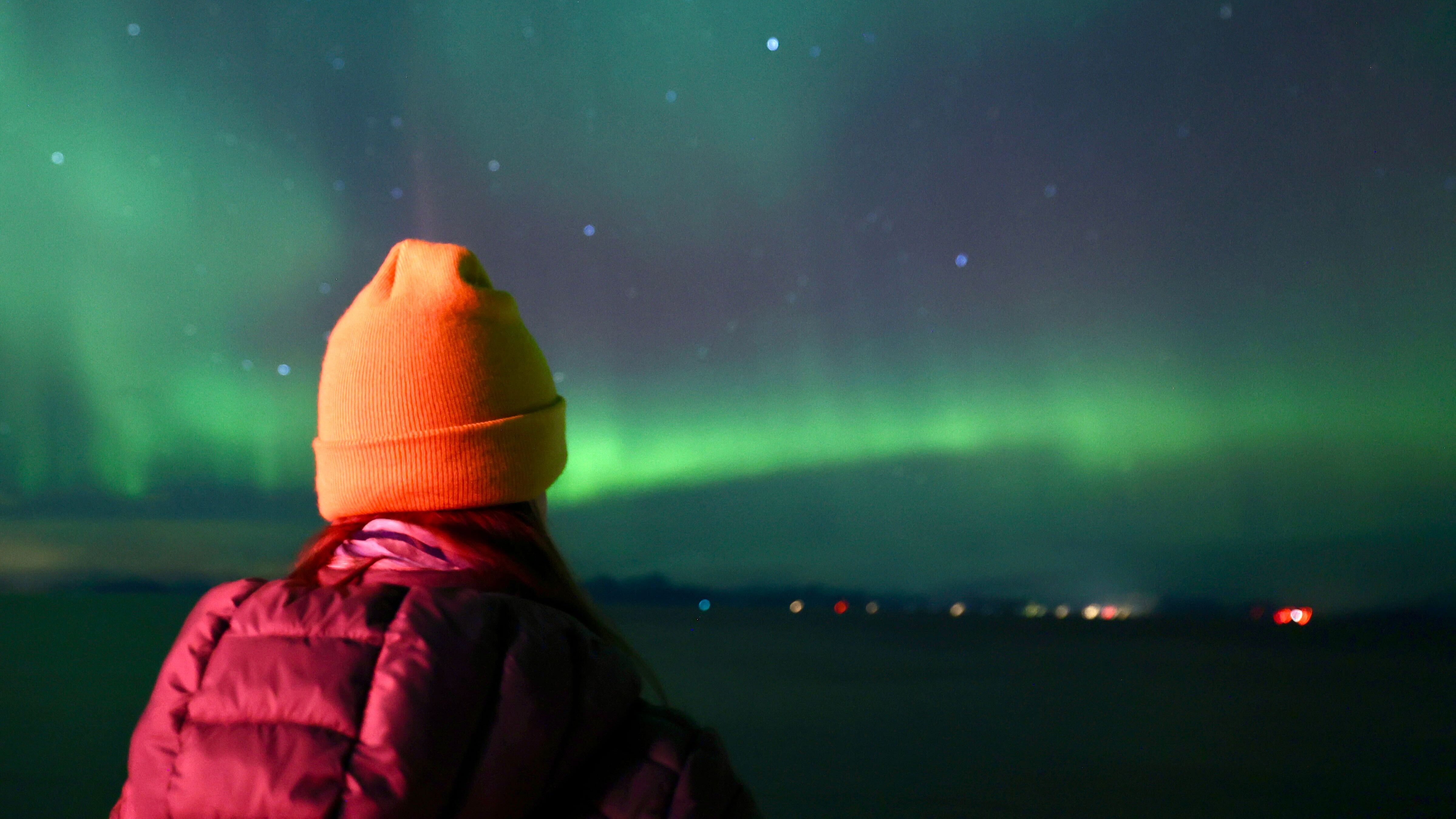 a woman wearing a yellow hat looks out to northern lights stretching across a starry sky.