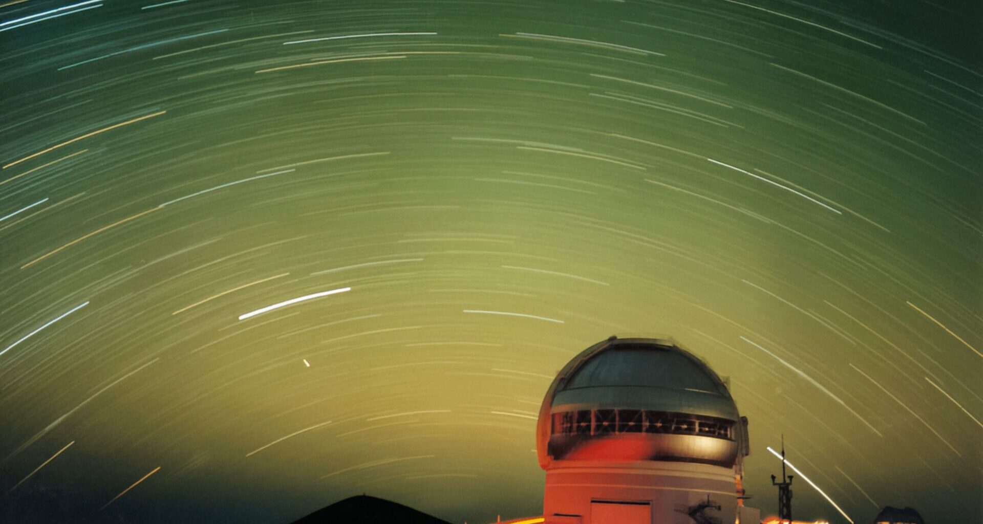 A series of white streaks move in a circular fashion in a green night sky all over the lit up dome of an observatory at the bottom right of the image.