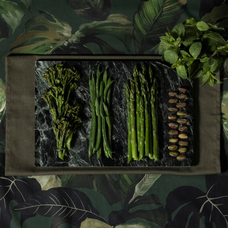 Broccolini, green beans, asparagus, mint leaves, and rows of pistachios are neatly arranged on a dark marble tray atop a green botanical-patterned tablecloth.