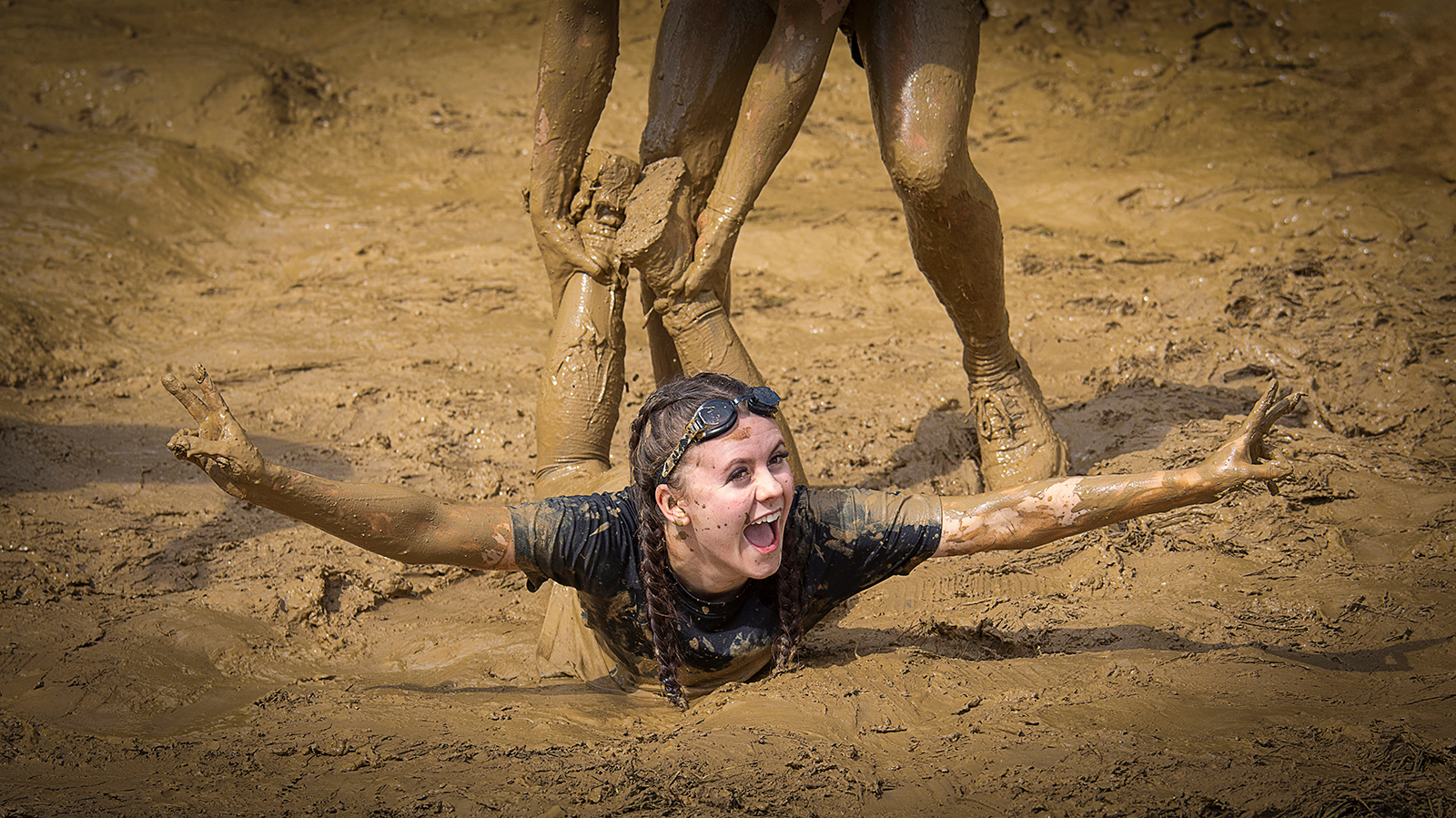 A person partially submerged in muddy ground, reaching out with arms while others assist them from above, surrounded by a muddy landscape