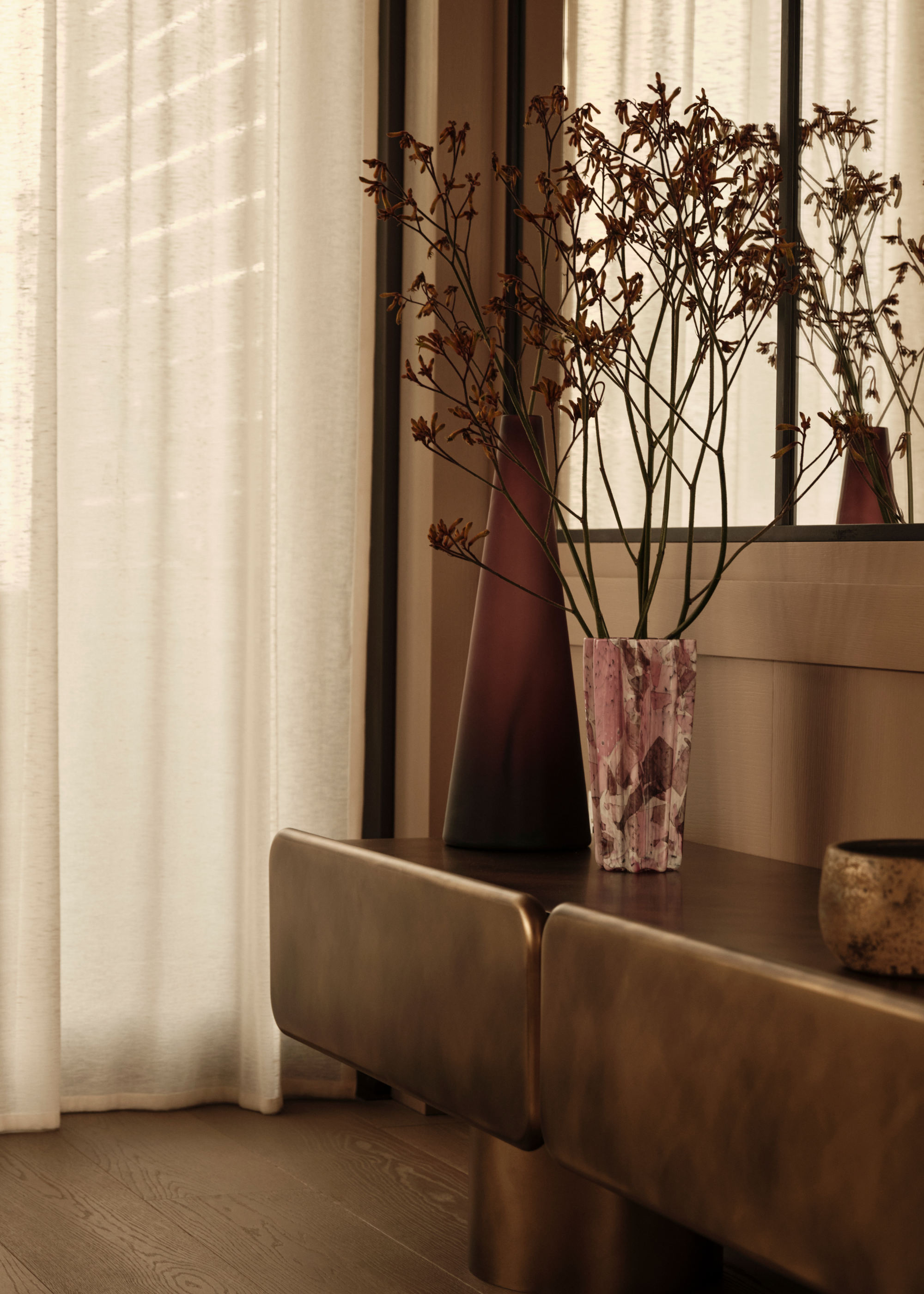 A dark brushed gold credenza with some decorative objects and a vase of dried foliage with sheer cream curtains pulled across the window