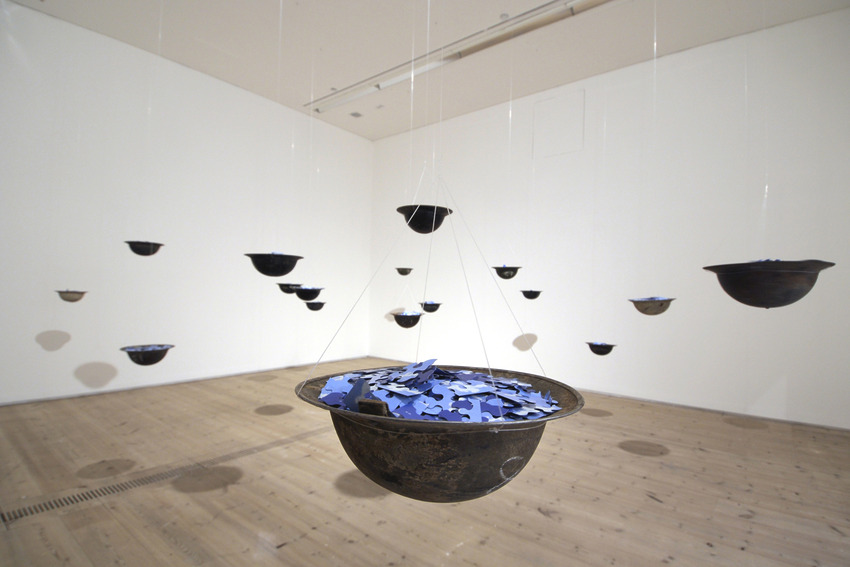 Art installation of suspended black bowls filled with blue pieces, hanging in a minimalist white gallery space.