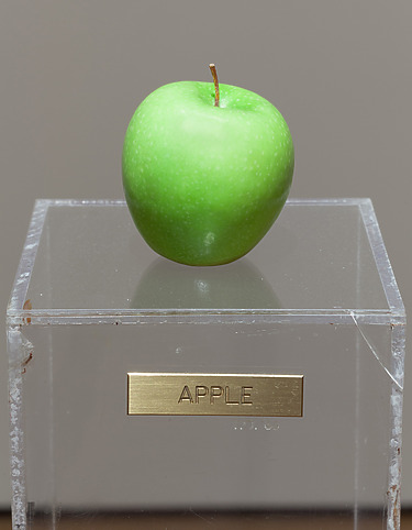 Green apple displayed on a clear pedestal with a brass label, set against a neutral background.