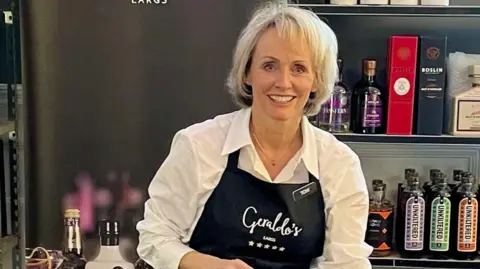 Toni Dawson A woman with blonde hair standing behind the counter of a shop, wearing a sales apron. She is smiling to the camera.