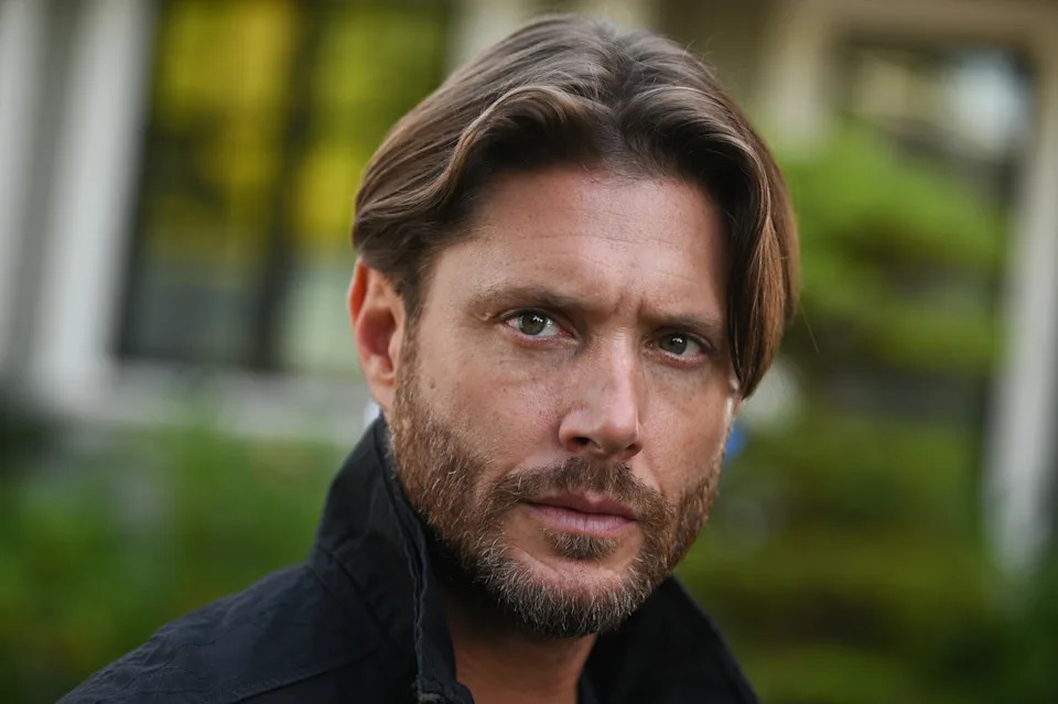 Jensen Ackles with a beard and medium-length hair wearing a dark jacket gazes intently at the camera, standing outdoors