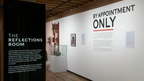London Museum The entrance to the Reflections Room with By Appointment Only printed on the white back wall. Art pieces can be seen in the background in glass cabinets.