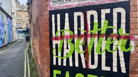 A black, green and white mural on the corner of an alleyway, which has a red cross painted roughly over it. 