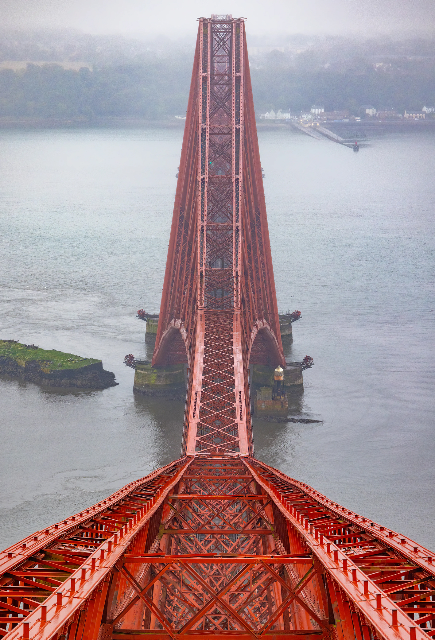 Red steel bridge arching over calm gray water on a foggy day, leading to a distant misty shoreline. The scene conveys a sense of serenity and grandeur