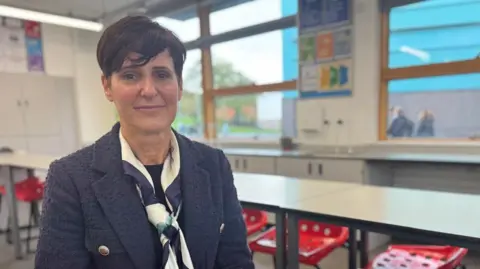 Becky Francis is seated at a table in a classroom wearing a dark textured jacket and a patterned scarf. The room has white walls, large windows letting in natural light, and posters with educational content on the wall. There are red plastic chairs with holes in the seat arranged around white tables.