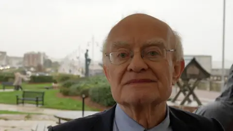 Jamie Niblock/BBC A head and shoulders image of Sir John Rutter. He is wearing glasses and a navy suit jacket over a light blue shirt. He is standing outside and looking into the camera. It is raining behind him. 